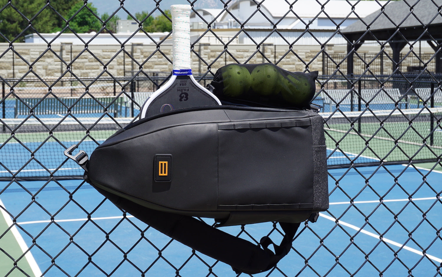 Black FORWRD Cross Court pickleball sling bag hanging on a chain-link fence with paddle and ball storage, one of the best pickleball bags and a versatile pickleball backpack alternative.