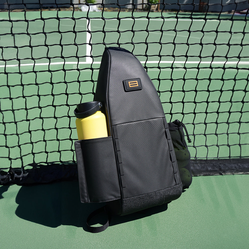 Black FORWRD Cross Court Pickleball Sling with single adjustable strap, side water bottle pocket, and mesh ball pocket, displayed on a green pickleball court by the net