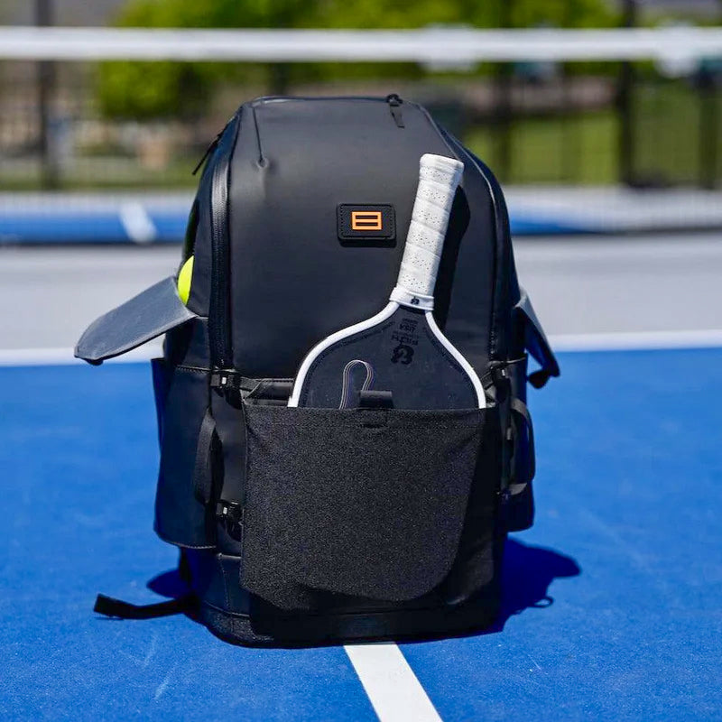 Black Court Caddy pickleball backpack on a bright blue court, featuring a white-gripped paddle in the front pocket—highlighting organized on-court gear storage.