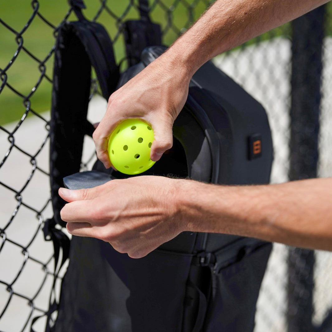 Person placing a bright green pickleball into the patented pending rapid pickleball system side pocket of a black Court Caddy pickleball backpack near a chain-link fence, highlighting convenient on-court gear access.