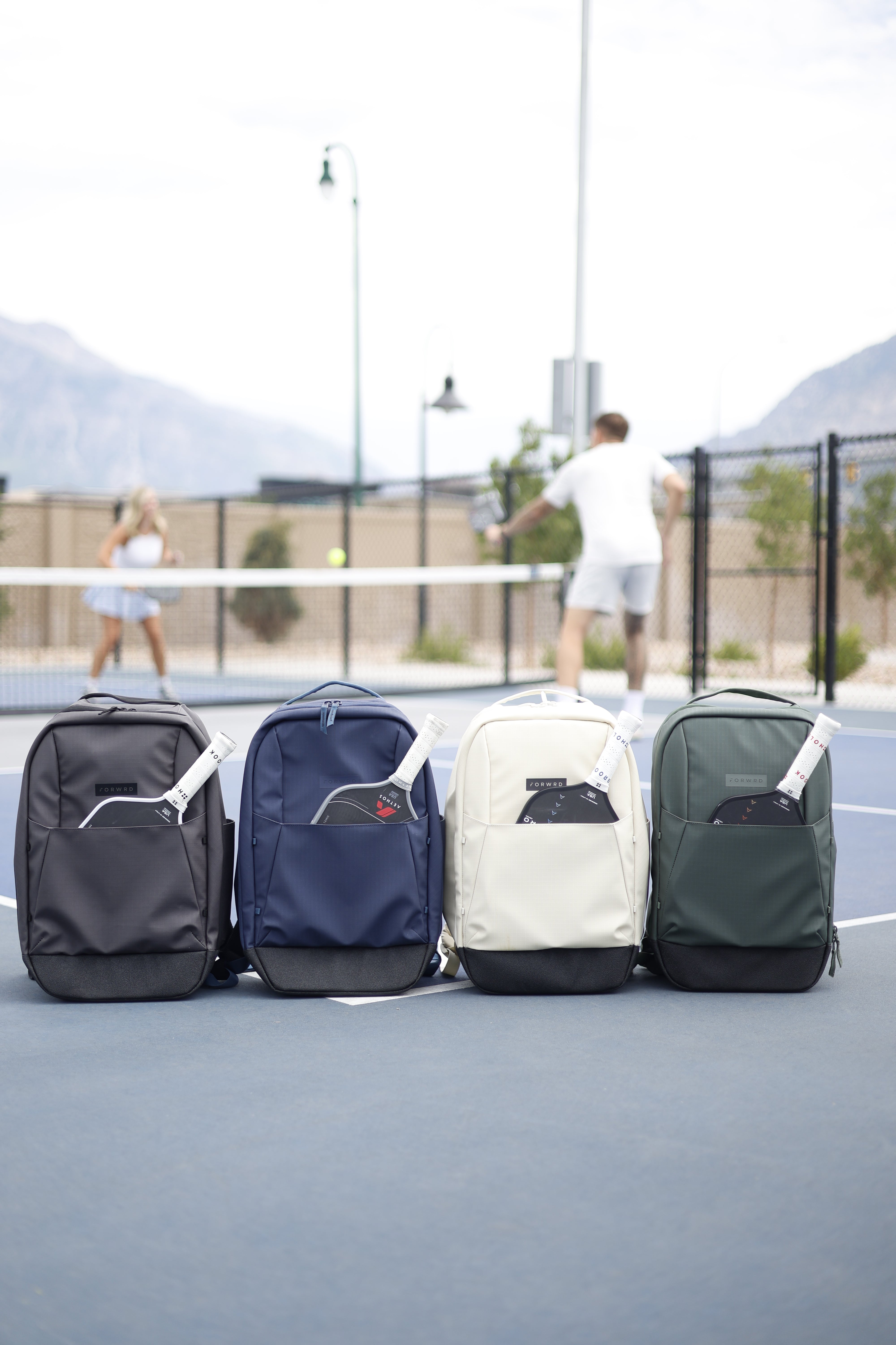 Four FORWRD pickleball bags with paddles displayed on court foreground, players rallying in background, premium gear setup
