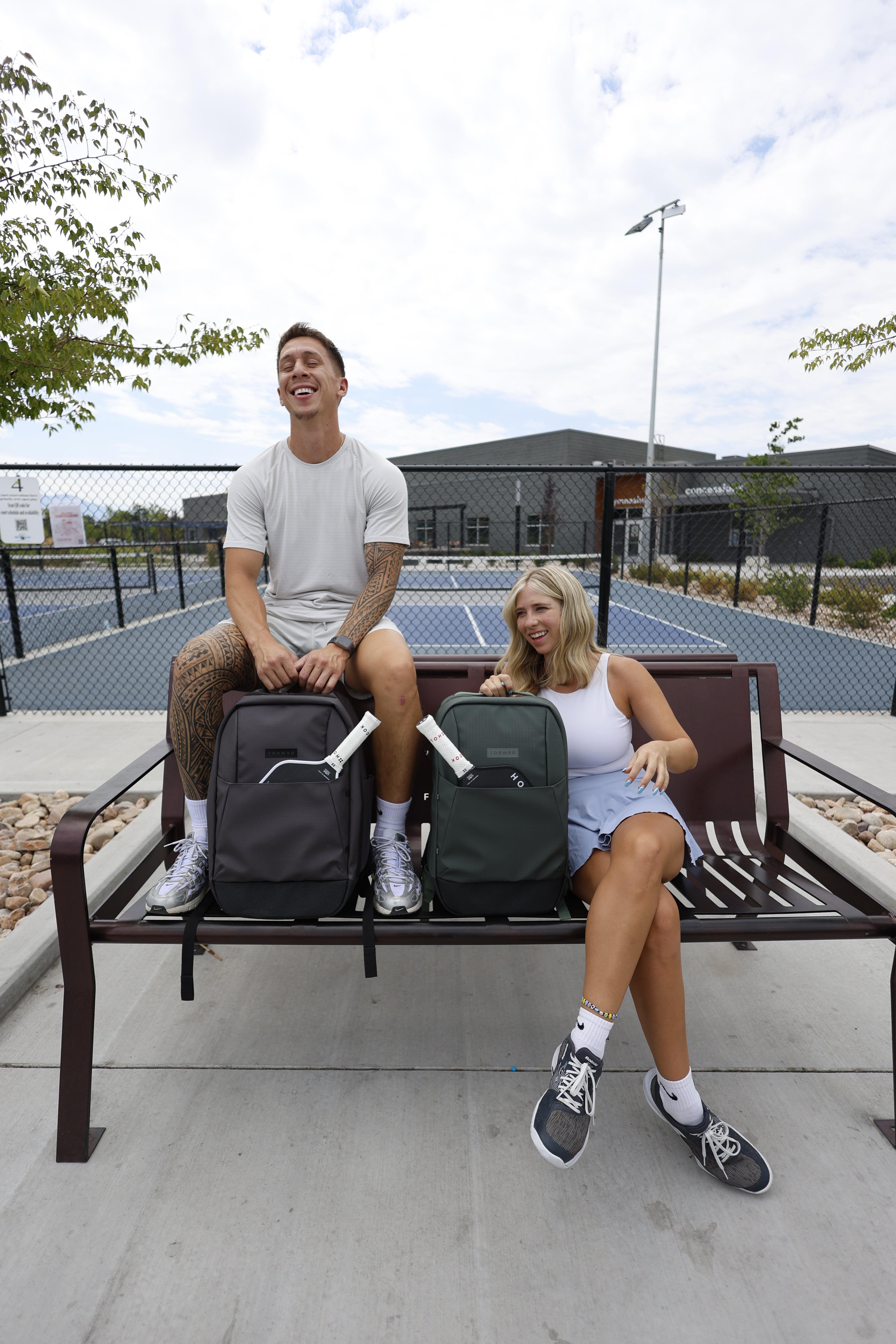 Man and woman sitting on bench outside pickleball court with charcoal and green FORWRD pickleball bags between them
