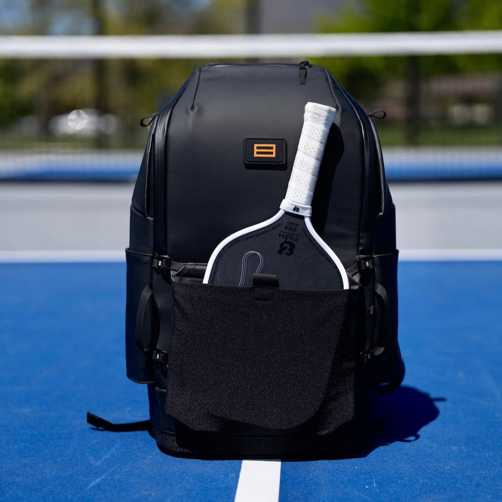 Black FORWRD Court Caddy pickleball backpack with modular add‑on paddle sleeve attachment holding a white‑gripped paddle in its front pocket, displayed on a vibrant blue court.