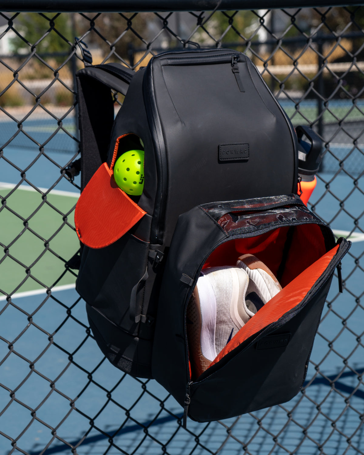 FORWRD Court Caddy pickleball backpack in the limited edition color black and orange, hanging on a black chain-link fence at a pickleball court