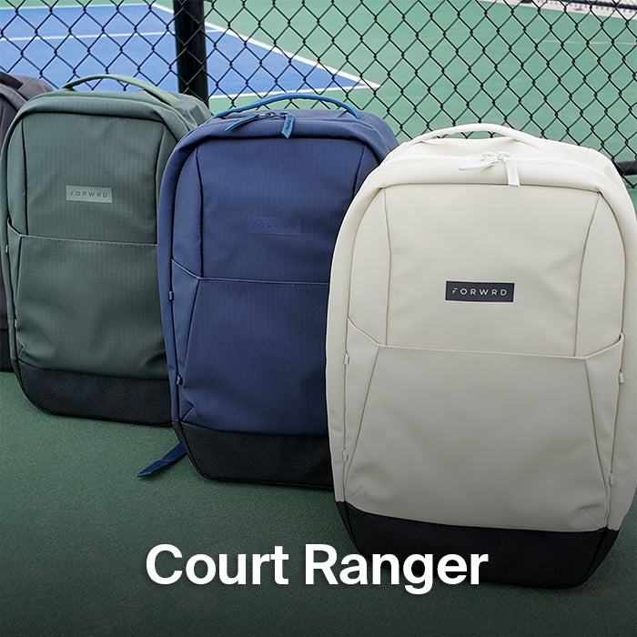 Three FORWRD Court Ranger pickleball backpacks in green, navy, and bone lined up on a court by a chain-link fence, showcasing color options for organized on-court gear storage.