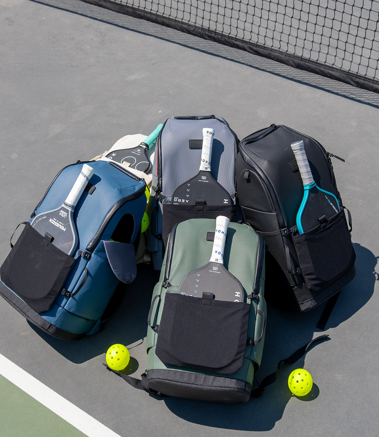 Assorted FORWRD Court Caddy pickleball bags on outdoor court with paddles and pickleballs in front of the net
